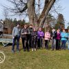 Na zdjęciu 11 osób z kijkami do nordic walking. Osoby pozują stojąc pod drzewem przy Ośrodku Edukacji Mazurskiego Parku Krajobrazowego.